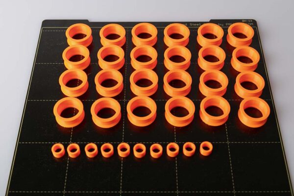 Print plate of the Prusa i3 MK3S on which there are 24 pieces of 3D printed pulleys and 10 pieces of 3D printed spacers. All parts are printed in orange PETG filament.