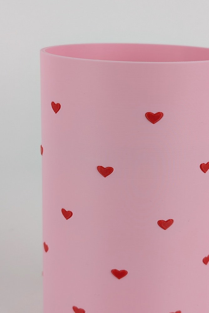 3D printed vase with a flower insert, printed in pink filament, featuring a design with a flower disc, where the hearts are red and embossed.