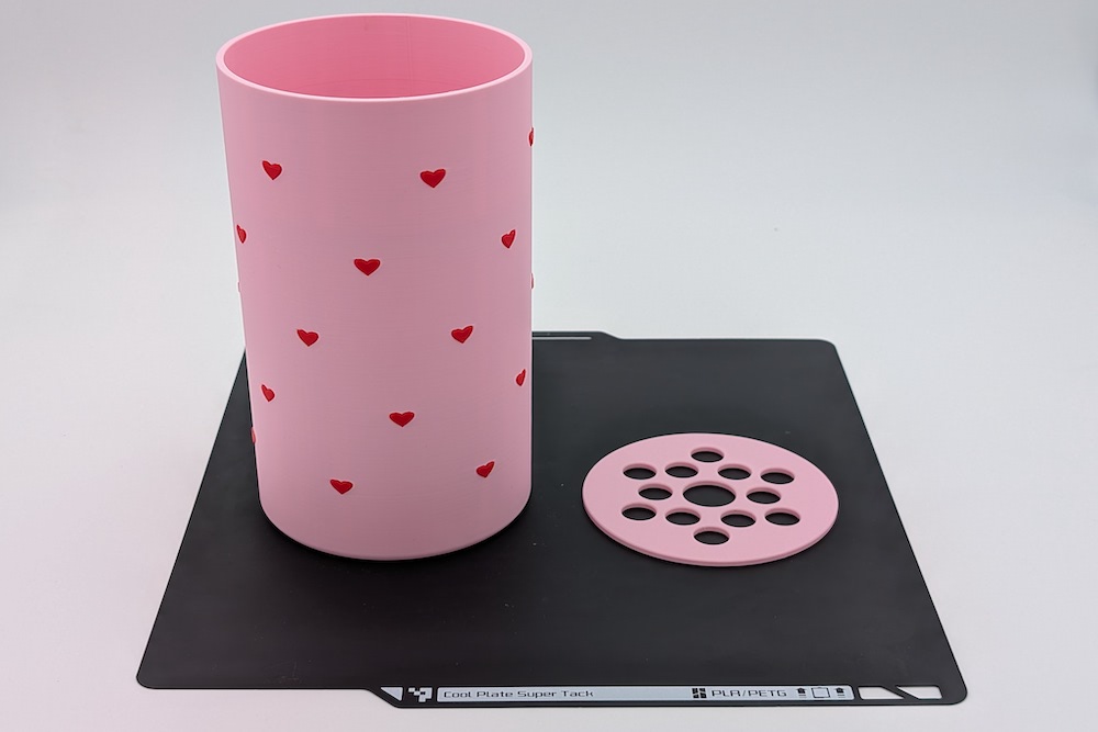 3D printed vase with a flower disc on the build plate, printed in pink filament, showcasing the flower disc design, where the hearts are printed in red and fully detailed.
