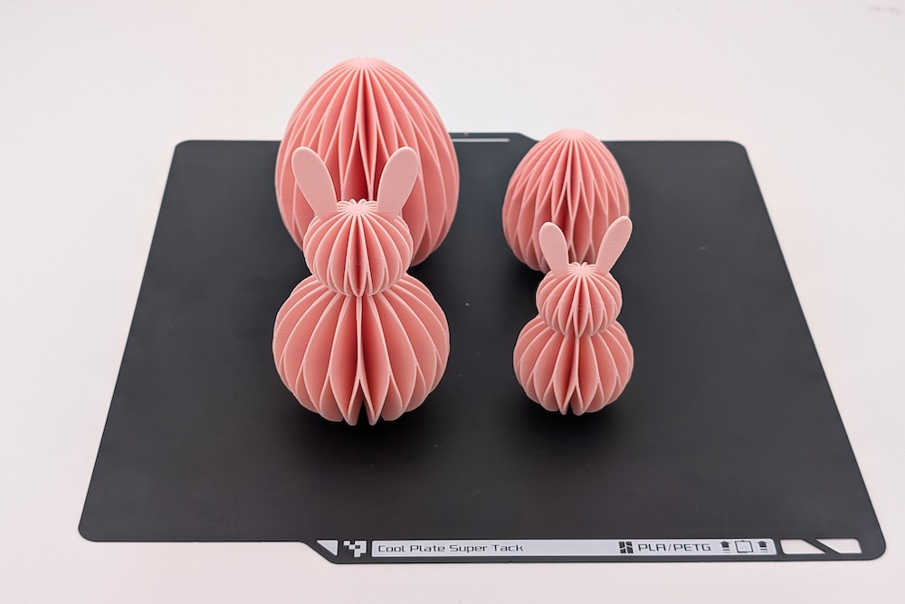 3D-printed honeycomb Easter decoration set (two bunnies and two eggs in various sizes) in pink, shown on a 3D printer build plate.
